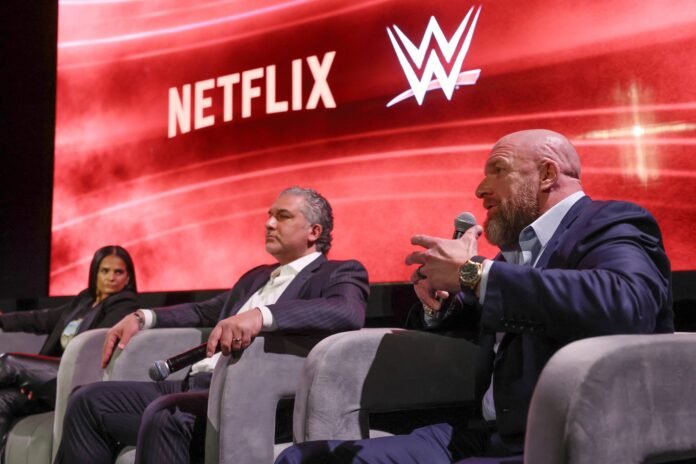 WWE Chief Content Officer Triple H speaks into a microphone sitting next to WWE President Nick Khan and Netflix Chief Content Officer Bela Bajaria on stage.