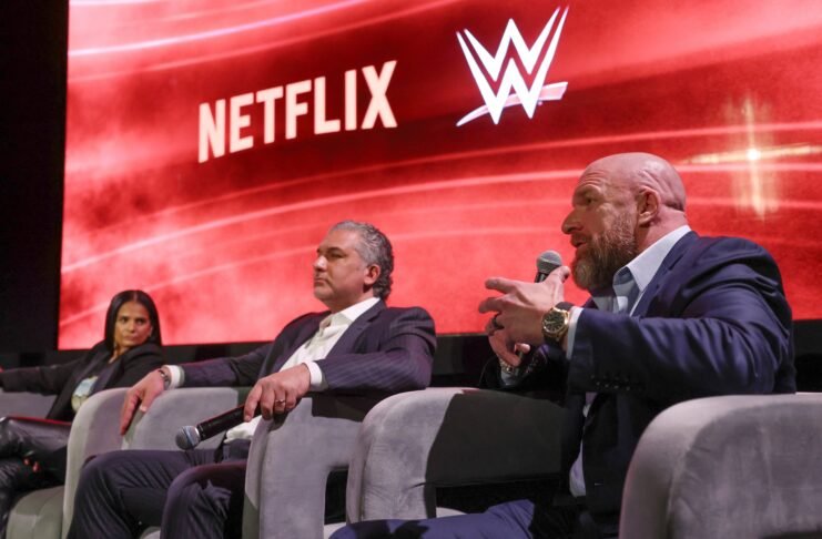 WWE Chief Content Officer Triple H speaks into a microphone sitting next to WWE President Nick Khan and Netflix Chief Content Officer Bela Bajaria on stage.