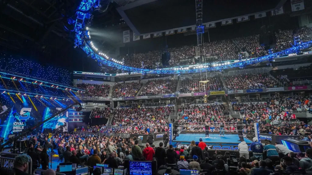 Wide shot of a packed arena hosting wwe smackdown, featuring a brightly lit wrestling ring in the center, stage lighting, graphics, and thousands of fans in attendance.