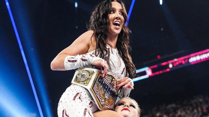 WWE Superstar Chelsea Green smiling enthusiastically while holding up her championship belt inside a wrestling arena.